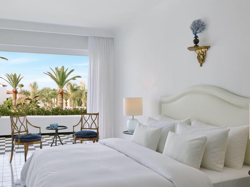 Světle zařízený hotelový pokoj s velkou manželskou postelí, posezením a výhledem na palmy.