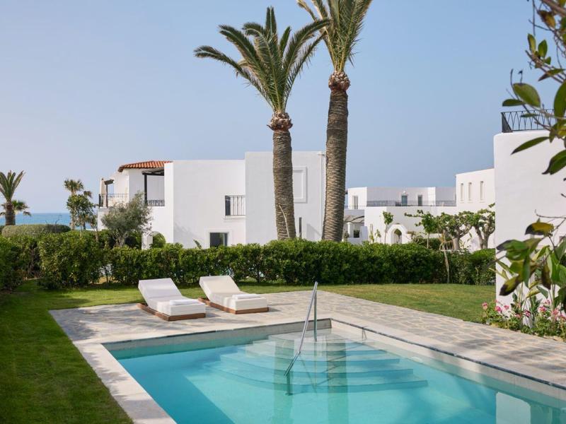 Bazén s lehátky, obklopený trávou a bílými budovami pod jasnou oblohou.