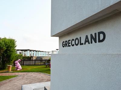 Außenansicht eines Gebäudes mit dem Schriftzug 'GRECOTAND' und einem pinken Hasen im Garten.