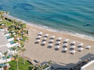 Strand mit Reihen von Sonnenschirmen, Palmen und angrenzenden Gebäuden neben ruhigem Meer.