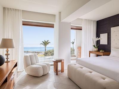 Helles Schlafzimmer mit kleinem Sofa, Holzmöbeln und Meerblick durch große Fenster.