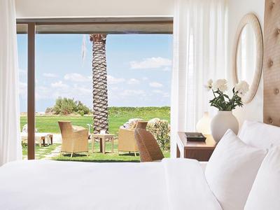 Helles Schlafzimmer mit großem Bett, Vorhängen, runden Spiegel und Blick auf Terrasse mit Stühlen.