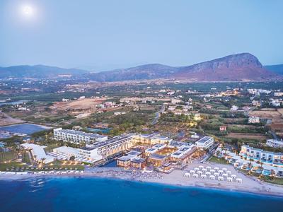 Küstenresort mit Strand, zahlreichen Gebäuden und Bergen im Hintergrund bei sonnigem Himmel.