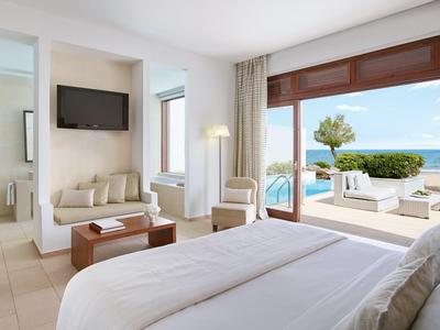 Helles Hotelzimmer mit großem Bett, Sitzecke und Blick auf Terrasse mit Pool und Meer.
