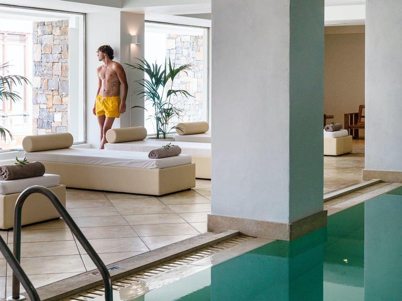 Innenraum mit grünem Pool, zwei Liegen und einer stehenden Person in gelber Badehose.