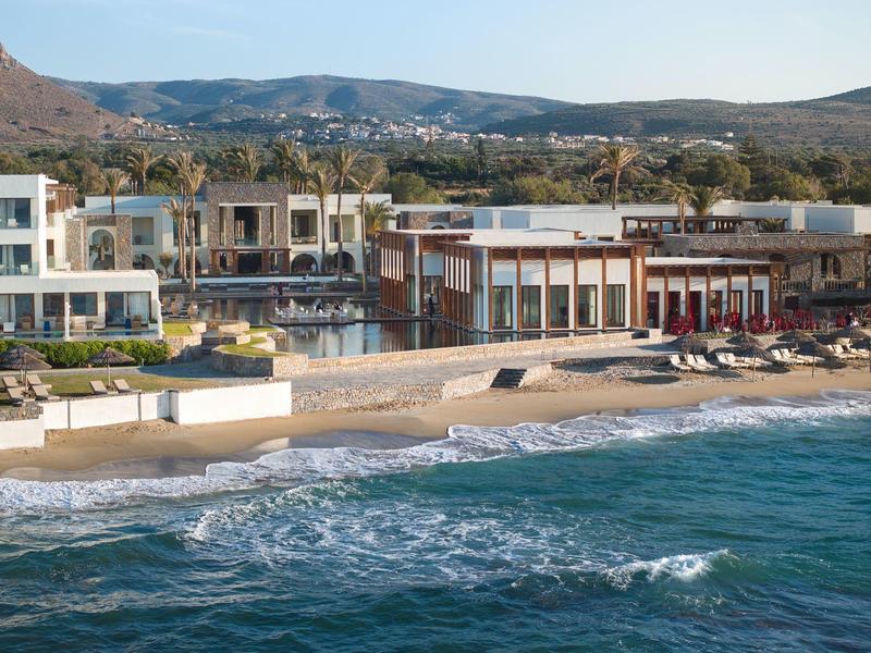 Strand mit Wellen, Liegen und weißen Gebäuden vor bewaldeten Hügeln bei klarem Himmel.