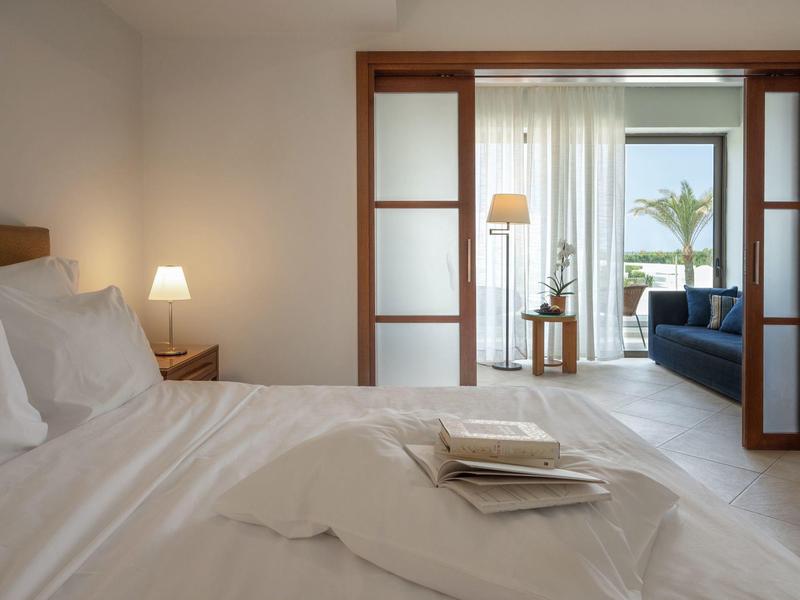 Helles Schlafzimmer mit weißer Bettwäsche, Holzrahmen, Lampe und Blick auf Balkon mit Palme und Sofa.