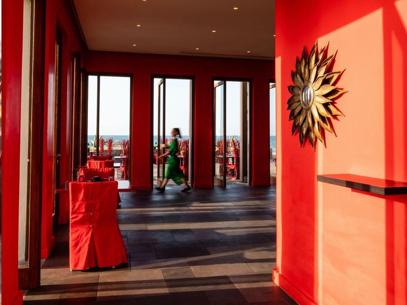 Roter Raum mit großen Fenstern, rotem Stuhl, Sonnen-Wanddekoration und zwei Personen im Hintergrund.