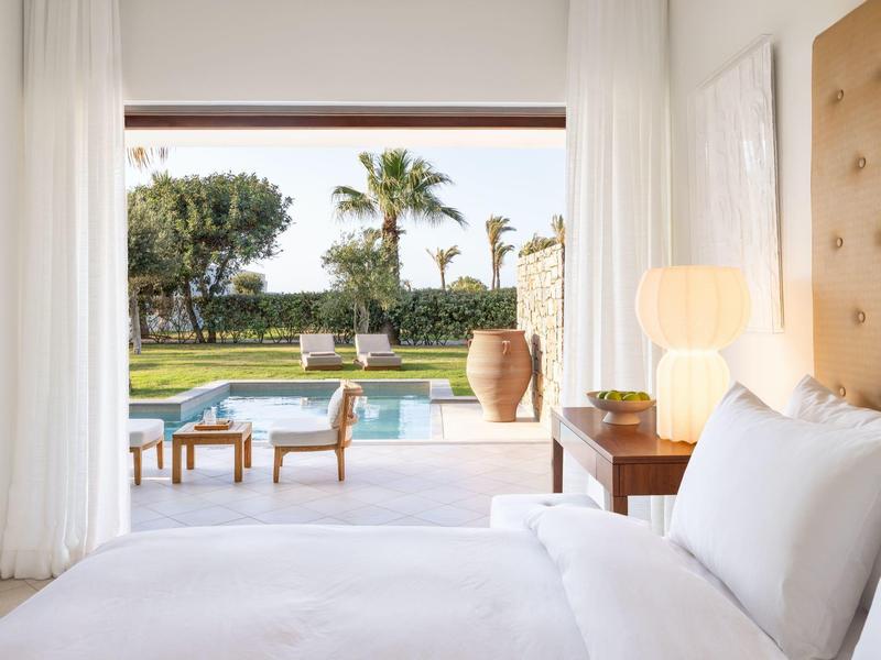 Hell eingerichtetes Schlafzimmer mit offenem Blick auf Terrasse, Pool und Garten mit Palmen.