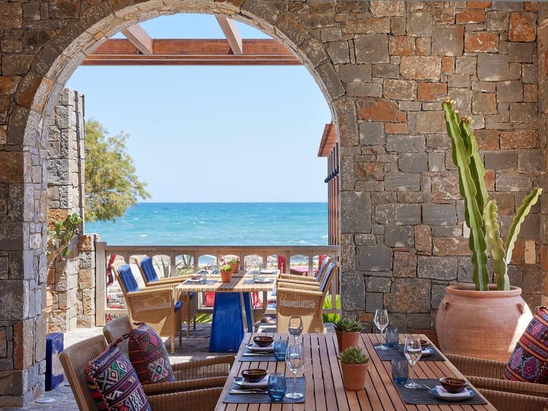 Terrasse mit Steinbogen, Esstisch, Stühlen und Blick aufs blaue Meer im Hintergrund.