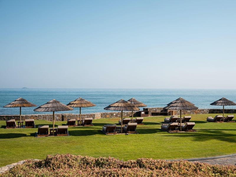 Grüne Wiese mit Holzbänken und Strohdächern vor ruhigem blauem Meer unter klarem Himmel.