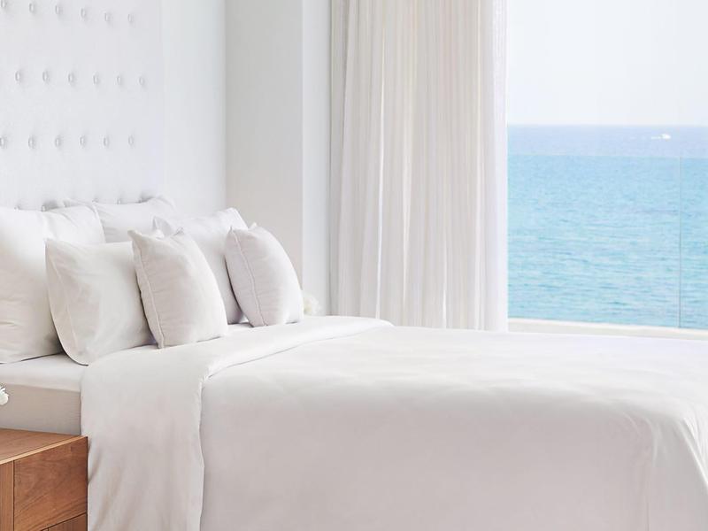 Helles Schlafzimmer mit weißem Bett, vielen Kissen und Blick auf das blaue Meer durch große Fenster.