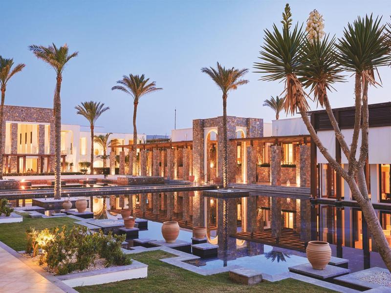 Moderner Gebäudekomplex mit Palmen, Wasserbecken und Holzelementen bei Abenddämmerung.