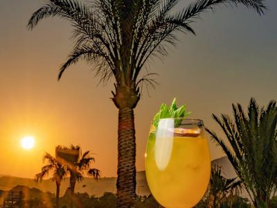 Sklenice koktejlu u bazénu při západu slunce s palmami v pozadí.