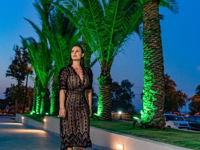 Žena v černých krajkových šatech kráčí osvětlenou cestou s palmami večer.