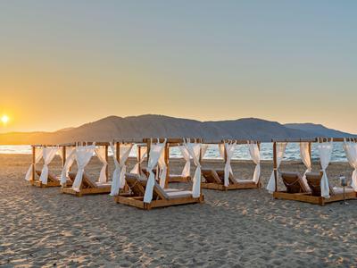 Pláž s bílými postelemi s baldachýnem a horami v pozadí při západu slunce.