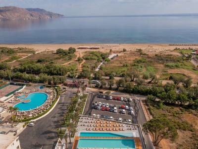Letecký pohled na hotel s bazénem, parkovištěm a výhledem na moře a pobřeží.