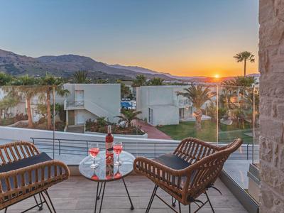 Dva židle a stůl s nápoji na balkóně při západu slunce s výhledem na moderní domy a hory.