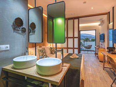 Moderní hotelový pokoj s otevřenou koupelnou, dvěma bílými umyvadly a výhledem na terasu a hory.