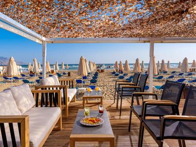 Jídelní prostor na pláži s stoly, židlemi a slunečníky u moře pod modrou oblohou.