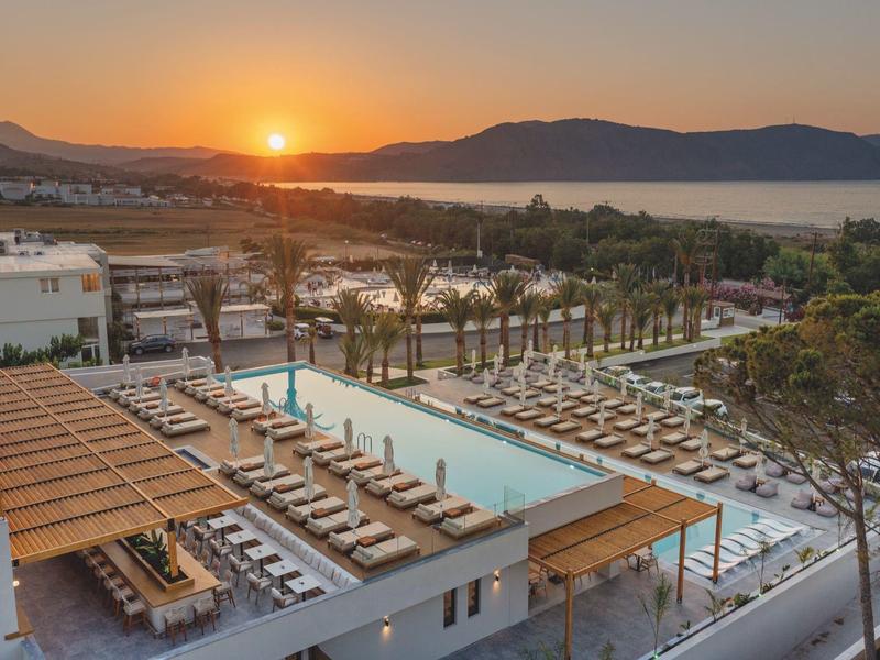 Západ slunce nad resortem s bazénem, lehátky a výhledem na moře a kopce.