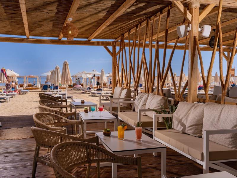 Plážový bar s útulným dřevěným posezením a výhledem na písek a slunečníky na pláži.