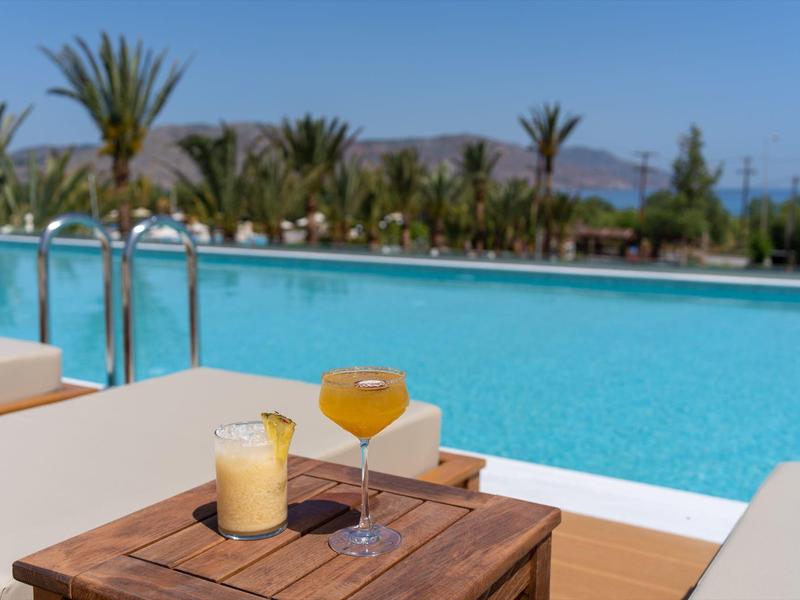 Dva barevné koktejly na dřevěném stole u bazénu s palmami a horami v pozadí.