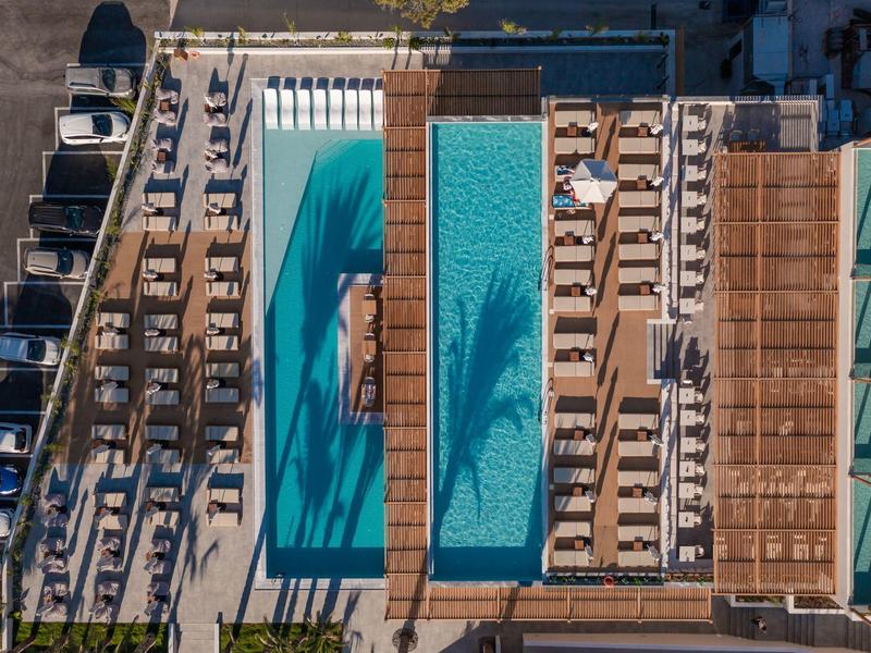 Letecký pohled na obdélníkový bazén s lehátky a zastíněnými pergolami vedle hotelu.