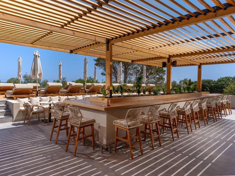 Moderní venkovní bar s dřevěnou pergolou, barovými židlemi a posezením za slunečného počasí.