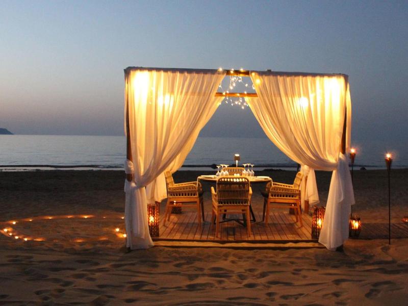 Romantická večeře pod osvětleným pavilonem na pláži při západu slunce.