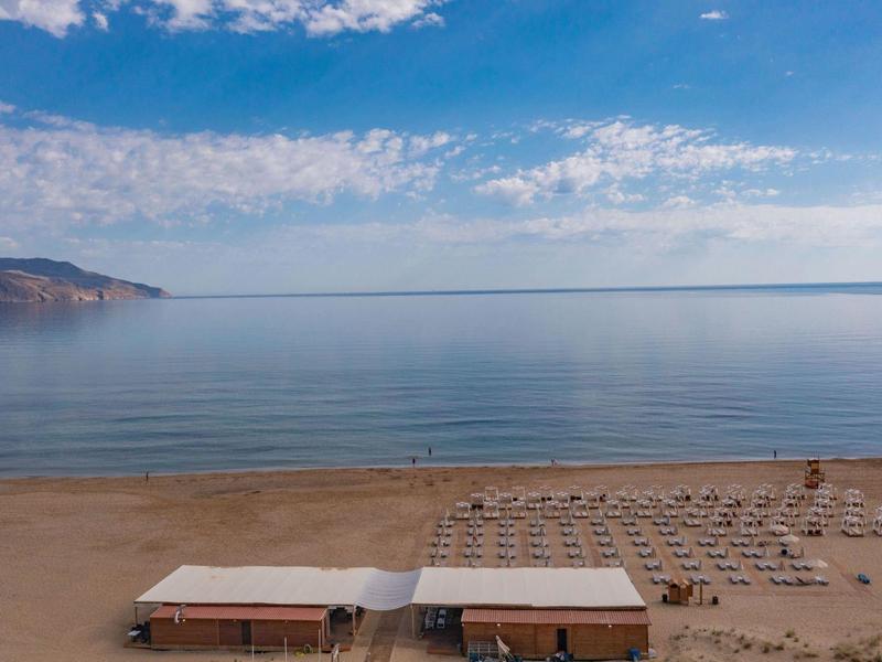 Prázdná písečná pláž s deštníky a dvěma malými budovami u klidného moře pod modrou oblohou.