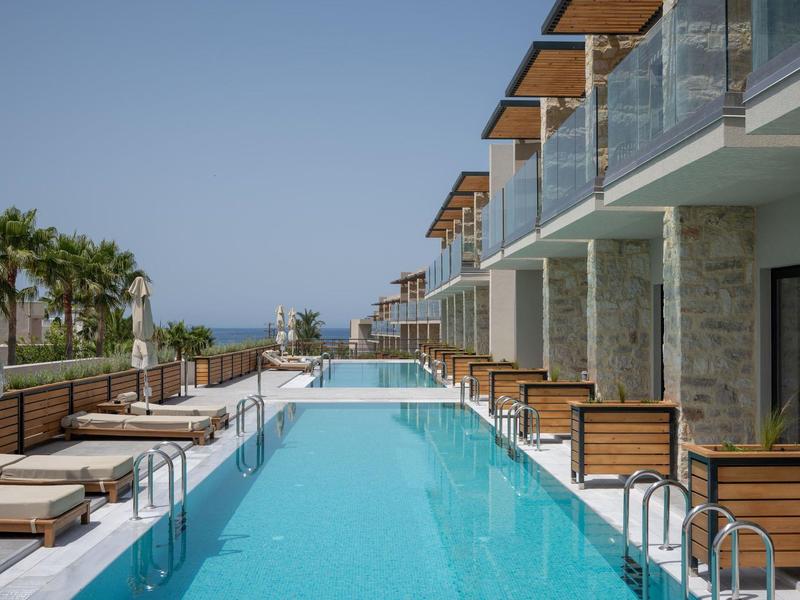 Moderní hotelový bazén s lehátky, skleněnými zábradlími a výhledem na moře v pozadí.