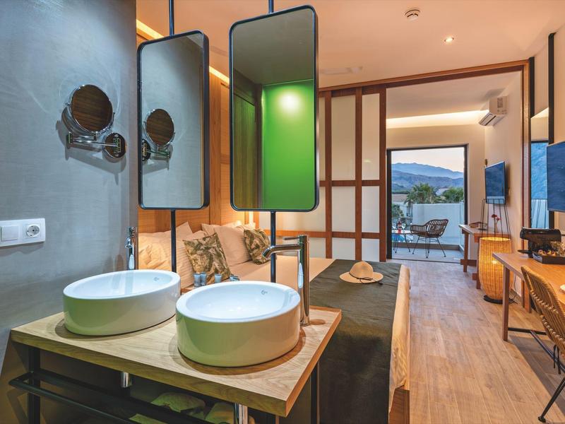 Moderní hotelový pokoj s otevřenou koupelnou, dvěma bílými umyvadly a výhledem na terasu a hory.