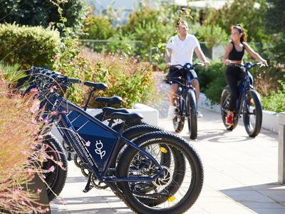 Deux personnes roulent à vélo électrique sur un chemin ensoleillé à côté de vélos électriques garés.