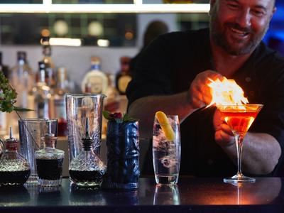 Barman podpalający jasnoczerwony koktajl w barze, na tle różne szklanki i butelki.