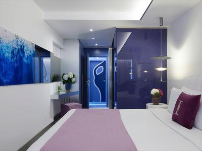 Nowoczesny pokój hotelowy z łóżkiem podwójnym, fioletowymi dekoracjami i dużymi drzwiami.