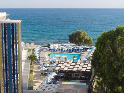 Vue sur une piscine d'hôtel avec parasols bleus, entourée de bâtiments et vue sur la mer.