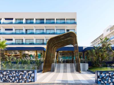 Entrée d'hôtel moderne avec bâtiment blanc, rambardes en verre et plantes tropicales le long du chemin.