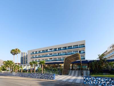 Hôtel moderne à plusieurs étages avec façade en verre, entouré de palmiers et ciel bleu.