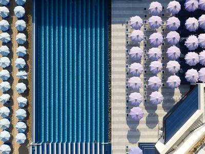 Vue aérienne d'une piscine avec des rangées de parasols bleus et violets dans un lieu de vacances hôtelier.