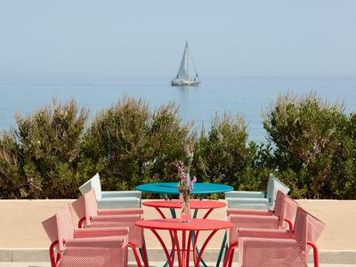 Terrasse avec chaises et tables rouges, vue sur la mer avec voilier et arbustes verts.