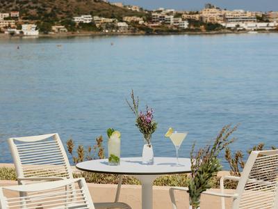 Une table ronde avec des boissons et des fleurs se trouve à côté de chaises blanches avec vue sur l'eau et un village.