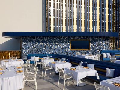 Espace extérieur moderne d'hôtel avec tables blanches, bancs bleus et bar carrelé de mosaïque.