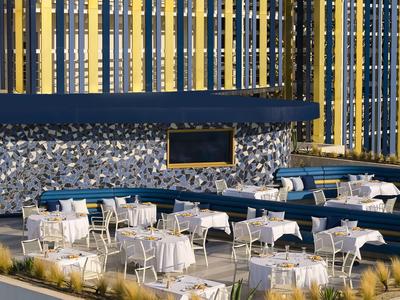 Espace extérieur moderne d'un hôtel avec des tables et chaises blanches devant une façade rayée jaune et bleue.