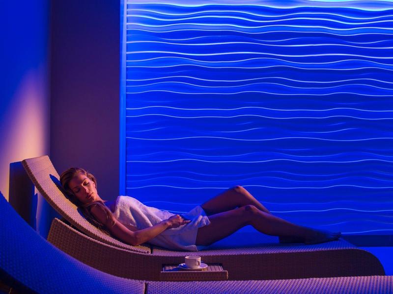 Kobieta relaksująca się na leżaku przy teksturowanej ścianie o niebieskim świetle, atmosfera spa.