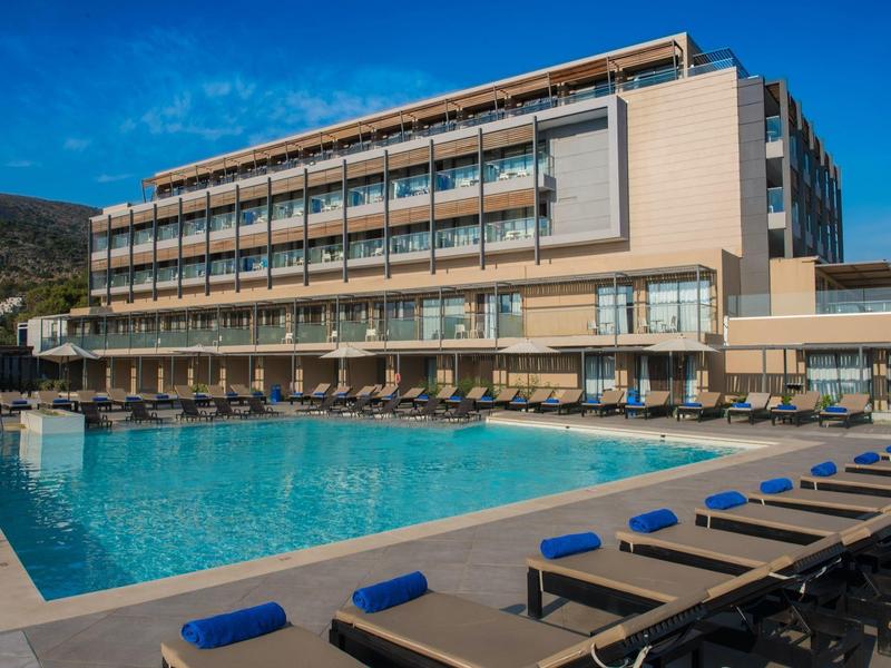 Nowoczesny hotel z dużym basenem na zewnątrz i wieloma leżakami pod bezchmurnym niebem.