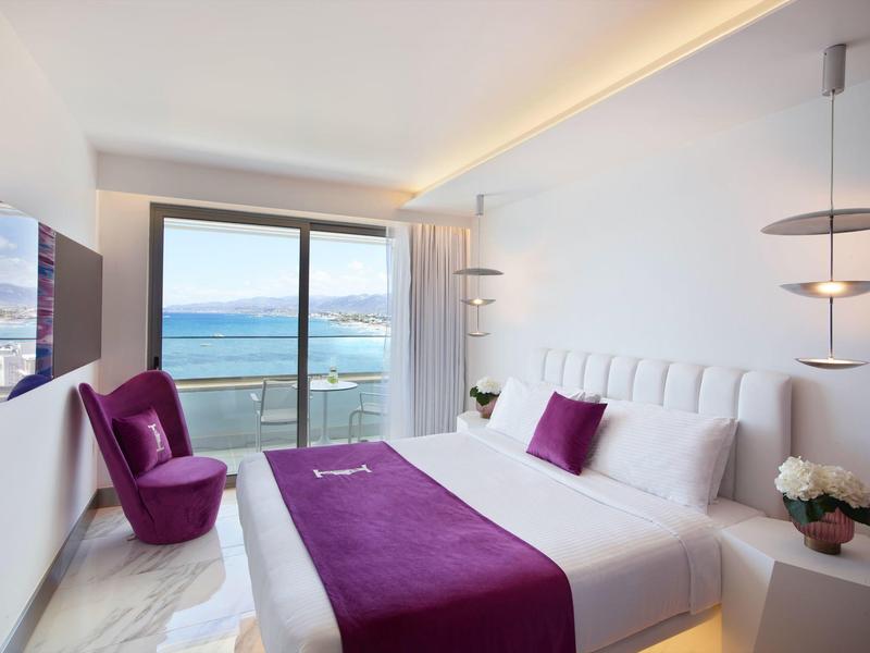 Nowoczesny pokój hotelowy z podwójnym łóżkiem, fioletowym fotelem i widokiem na morze z tarasu.