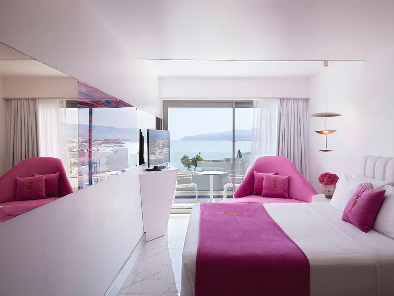 Nowoczesny pokój hotelowy z podwójnym łóżkiem, różowymi poduszkami i narzutą, duże okno z widokiem na morze.