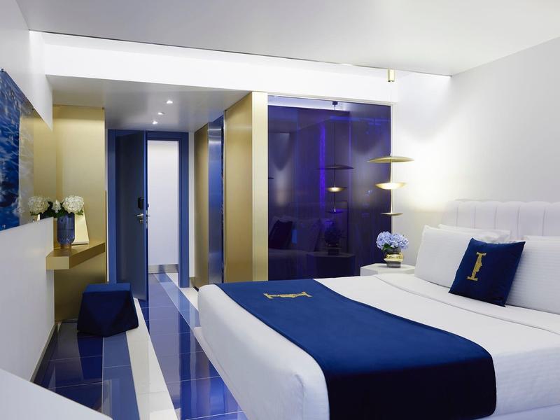 Nowoczesny pokój hotelowy z podwójnym łóżkiem, białymi i niebieskimi meblami, eleganckim oświetleniem.