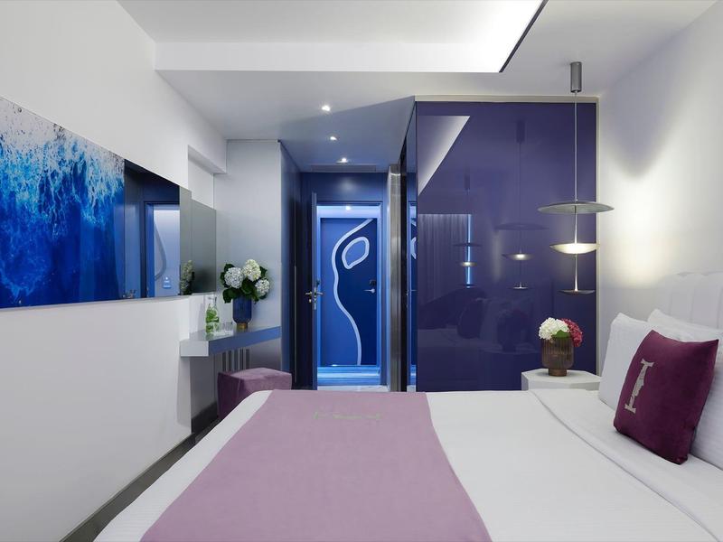 Nowoczesny pokój hotelowy z łóżkiem podwójnym, fioletowymi dekoracjami i dużymi drzwiami.